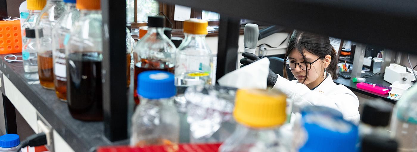 Woman st和ing behind a shelf full of colorful bottles in lab measures liquid with a pipette.